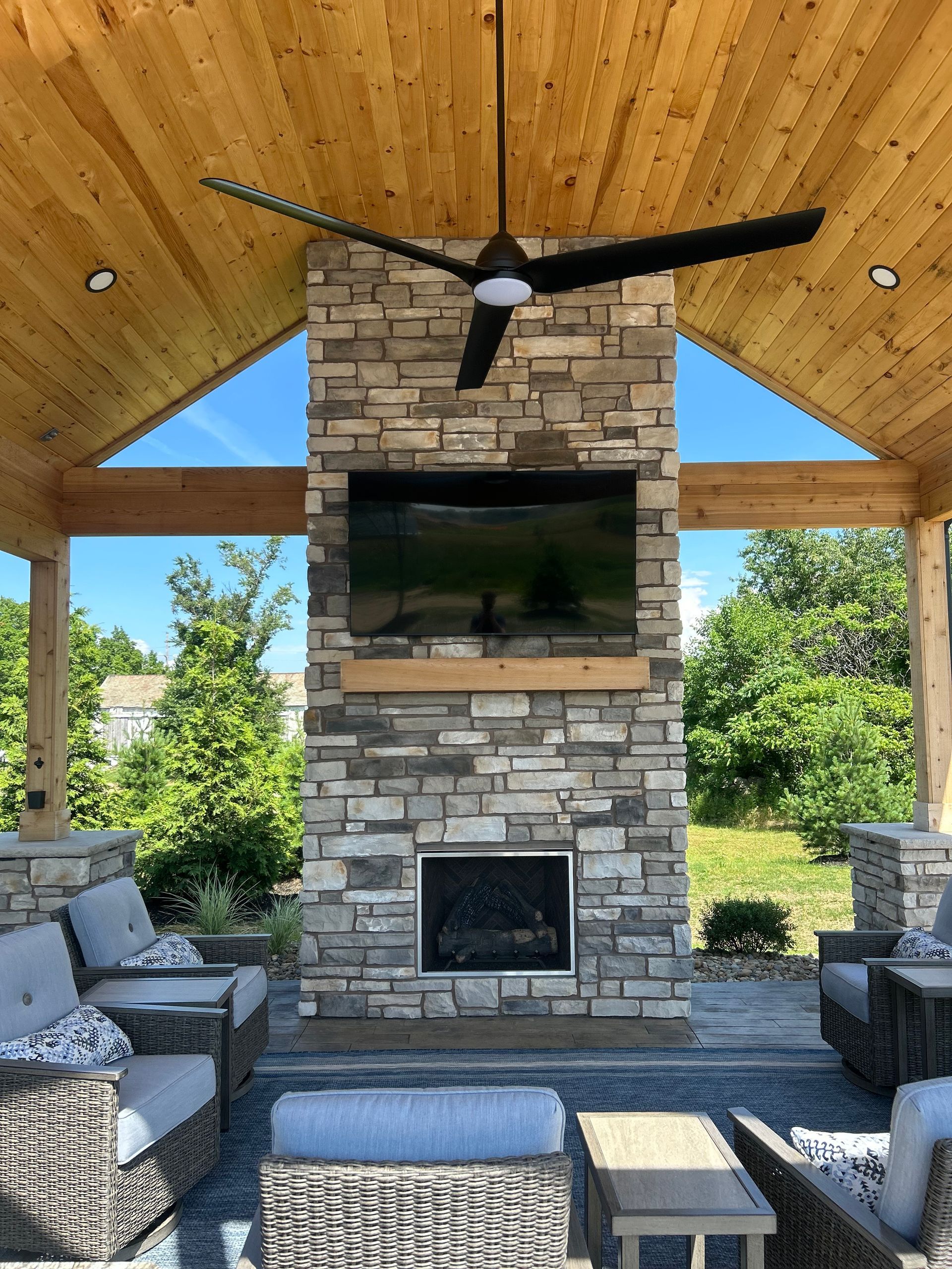 Outdoor patio with stone fireplace, TV, seating, and a ceiling fan.