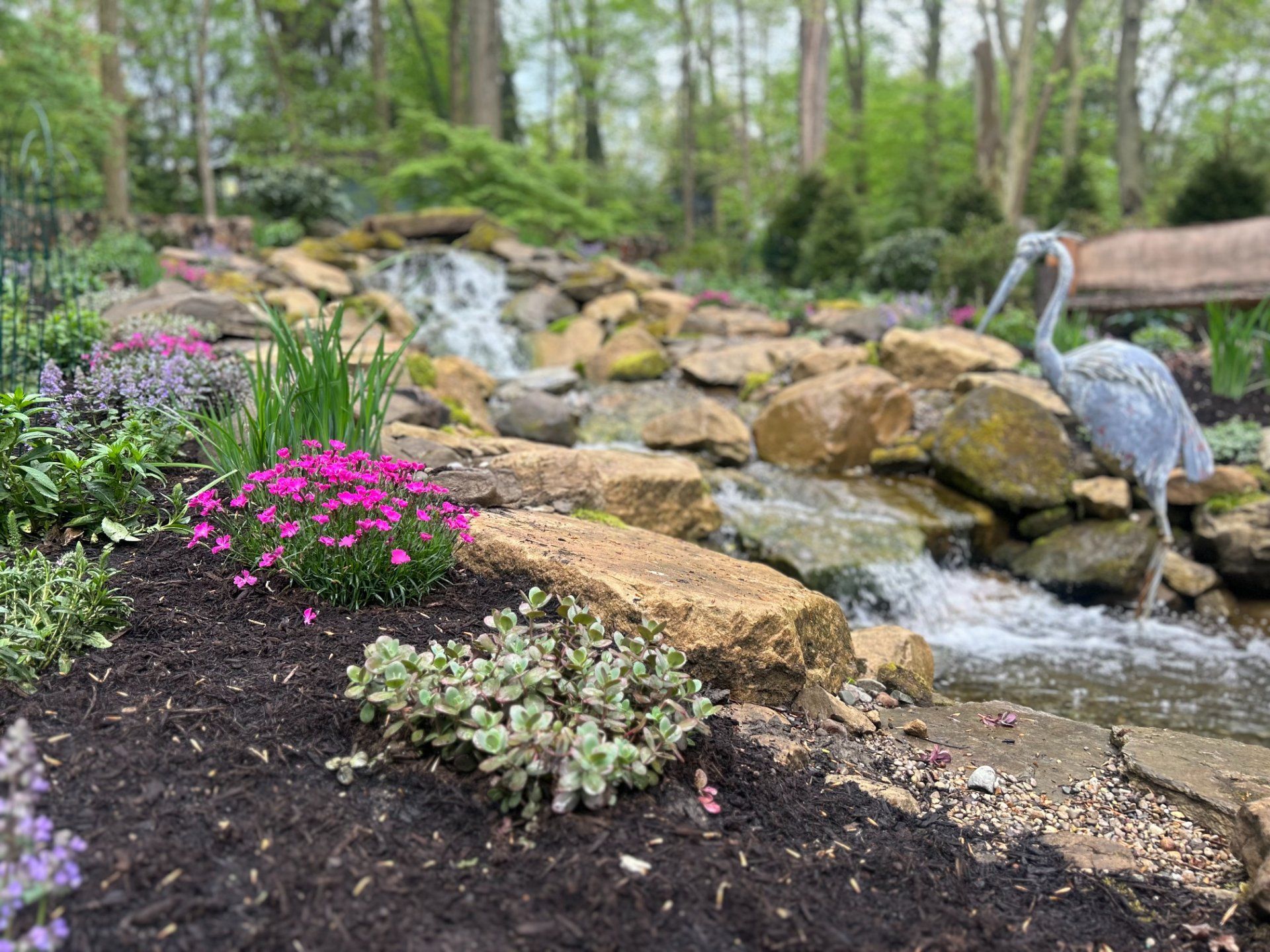Waterfall cascading over rocks in a garden; pink flowers and a heron statue near the water.