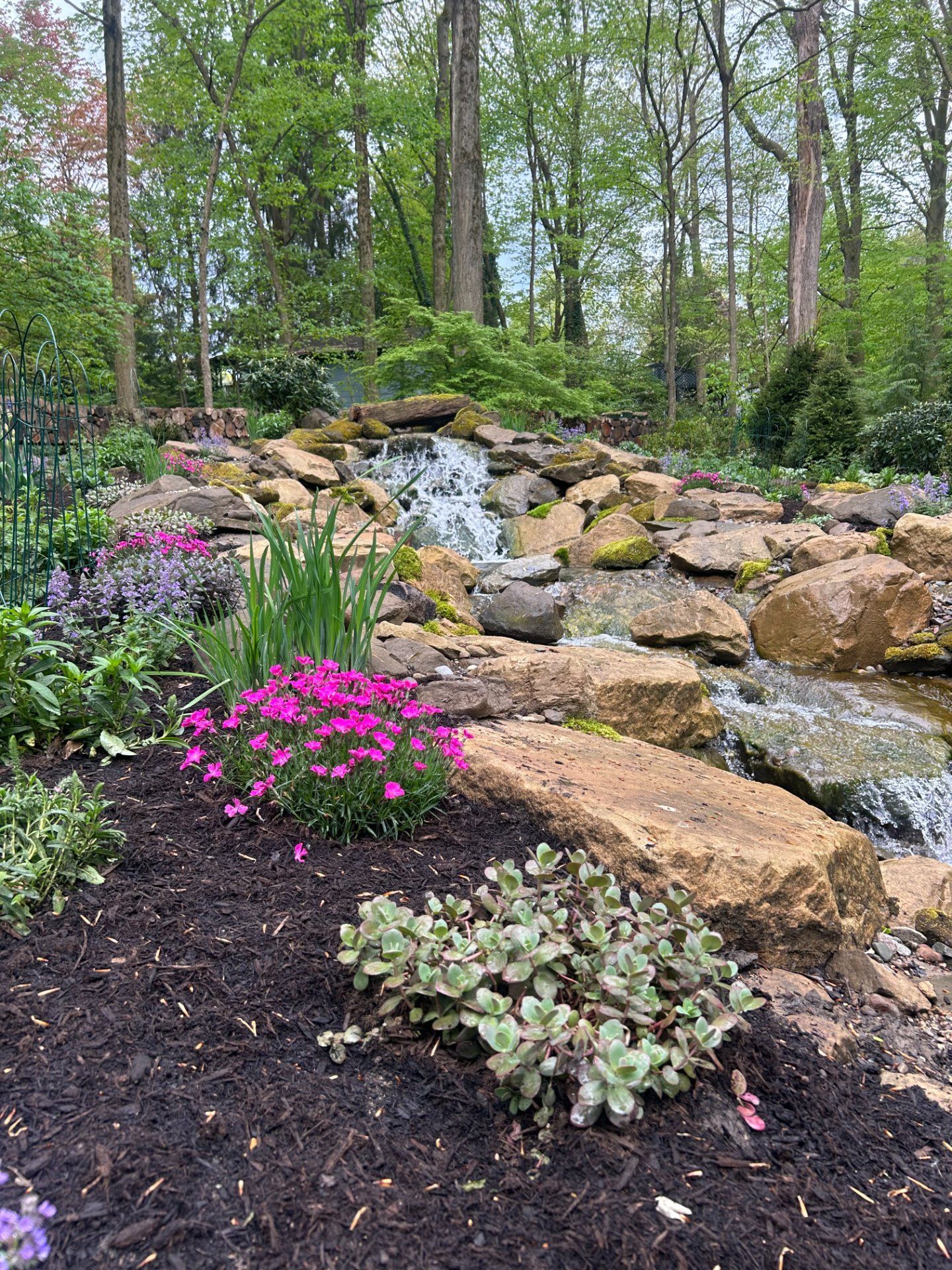 A waterfall cascades over rocks, surrounded by green trees and vibrant flowers, creating a serene garden scene.