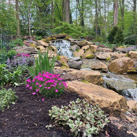 Waterfall cascading over rocks in a garden with pink flowers and lush greenery.