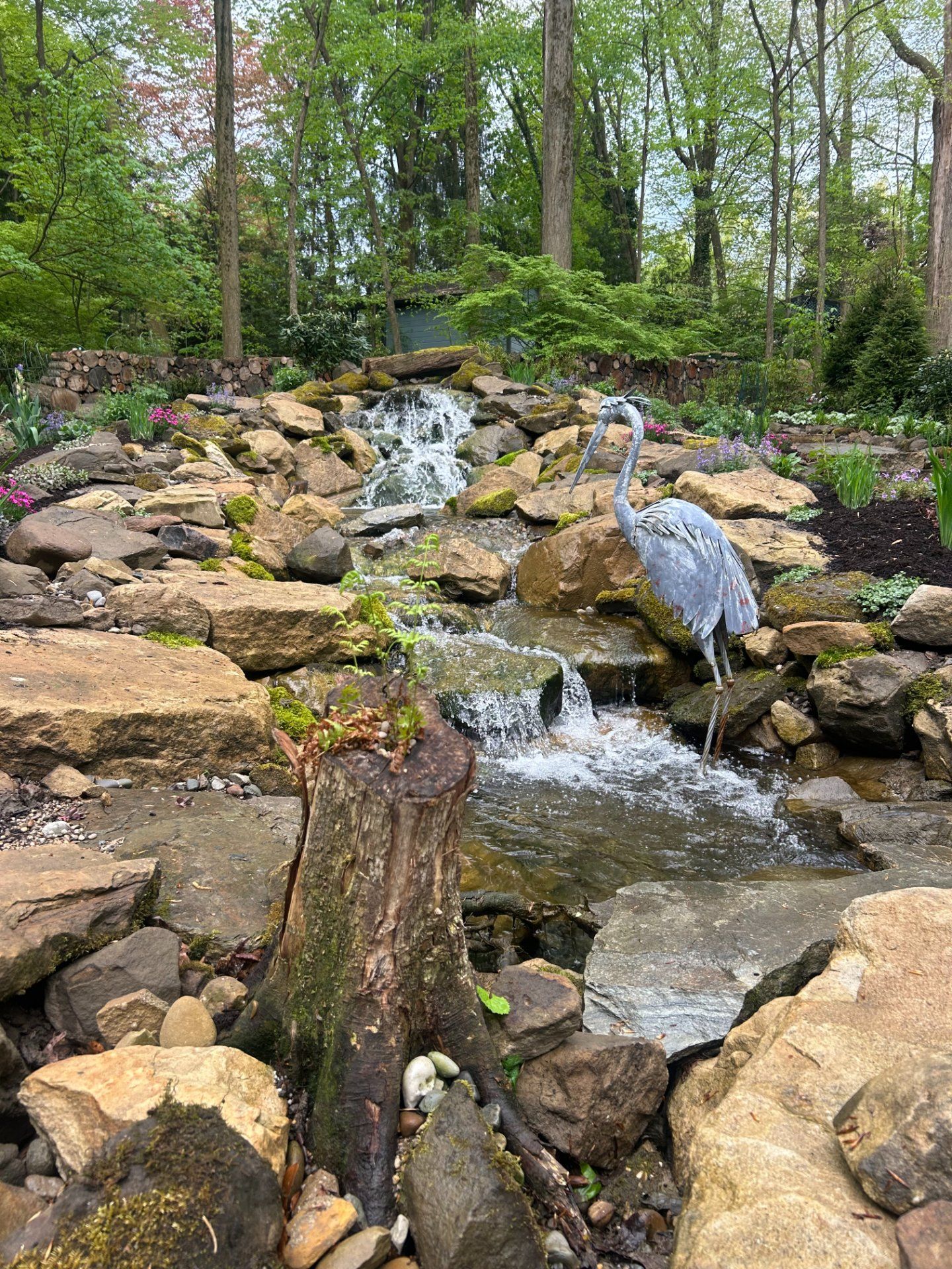 A stone waterfall with a blue heron statue, surrounded by greenery and rocks.