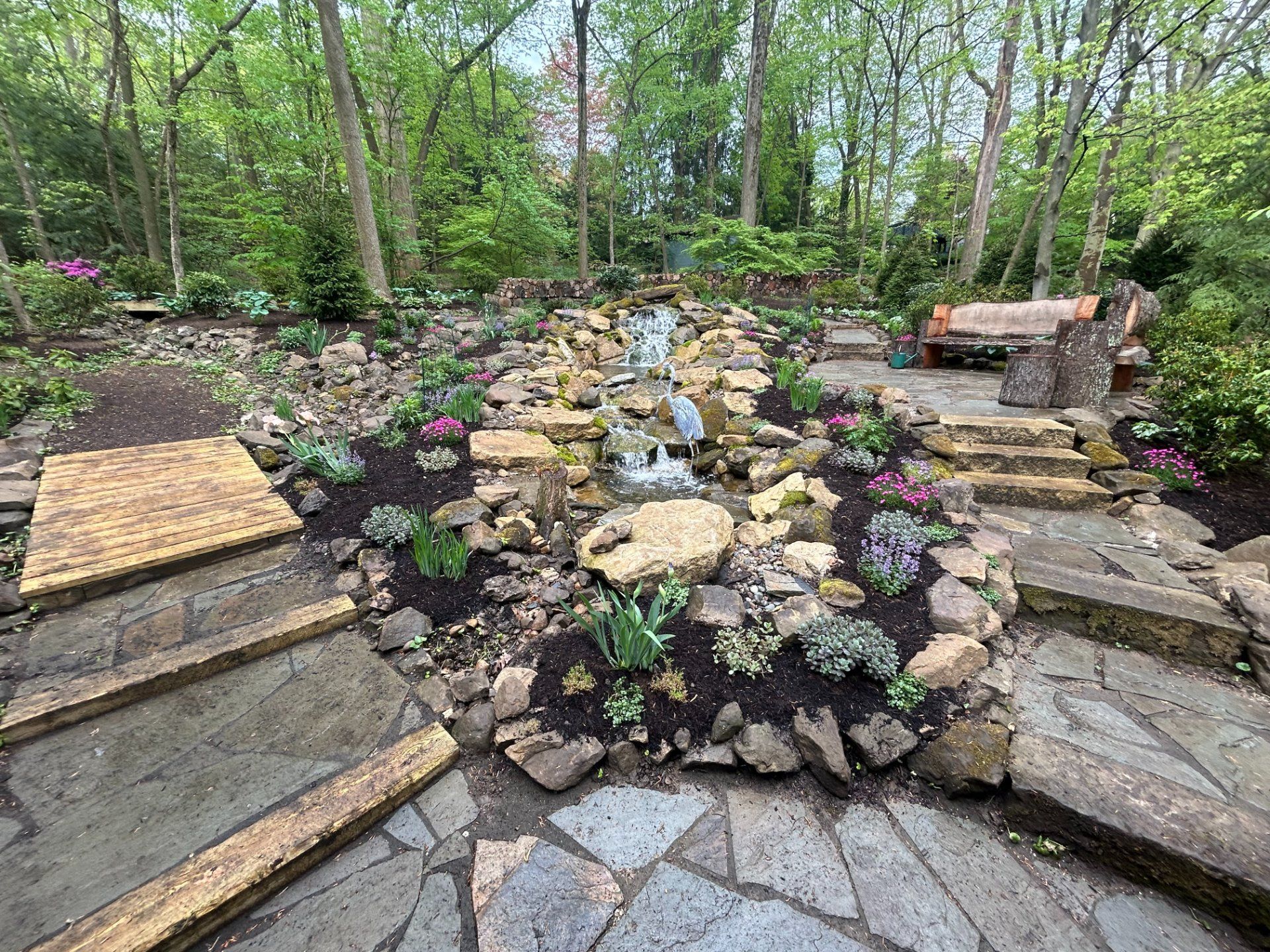 Stone waterfall feature in a garden, surrounded by plants and flagstone walkways, with a bench in the background.