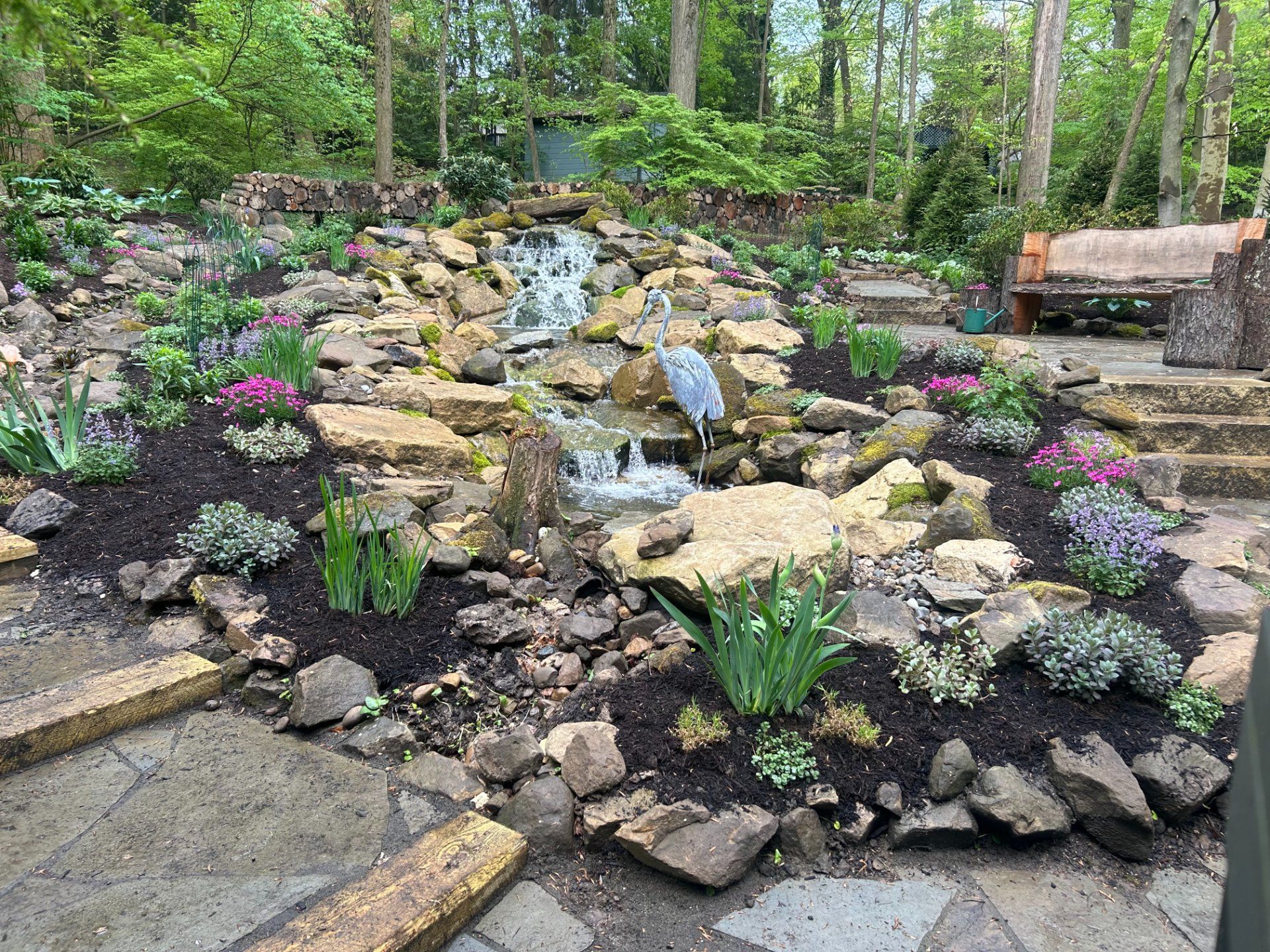 Waterfall in a landscaped yard with flowers and a wooden bench.