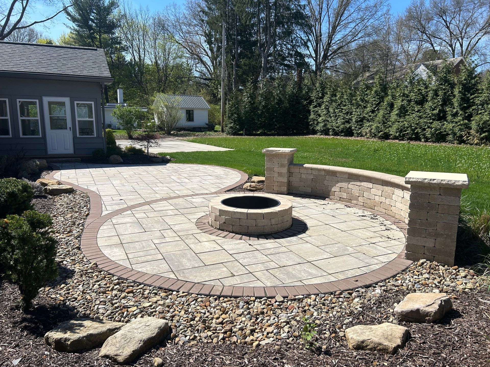 Stone patio with fire pit, curved wall, and landscaping in backyard.