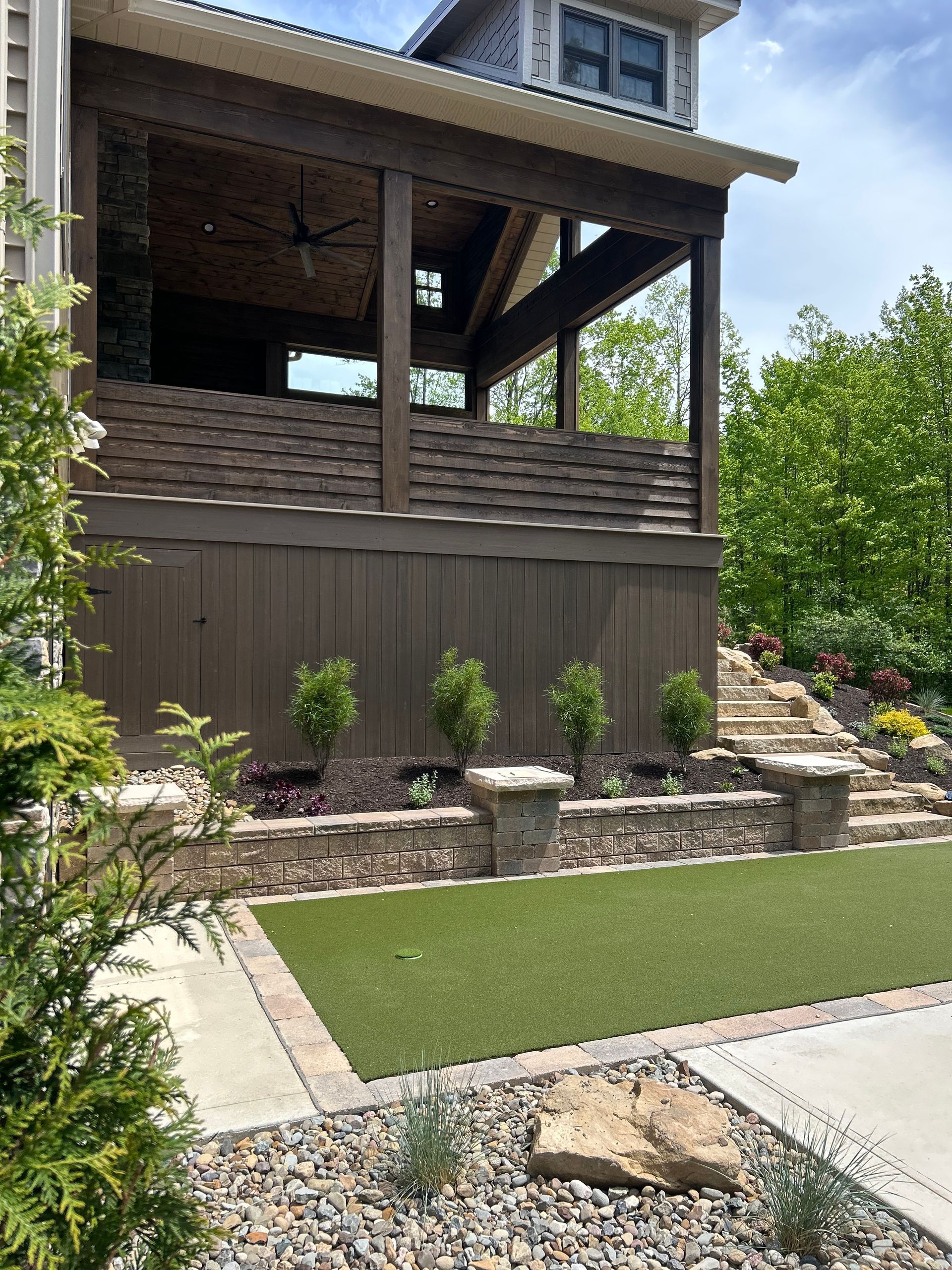 Covered outdoor living space with brown wood and stone retaining walls, small green shrubs, and green turf.