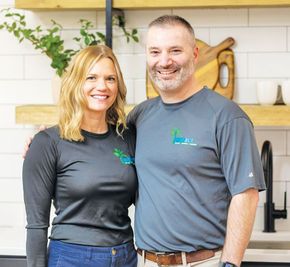 A smiling couple poses together in a kitchen. Both wear gray shirts with a logo.