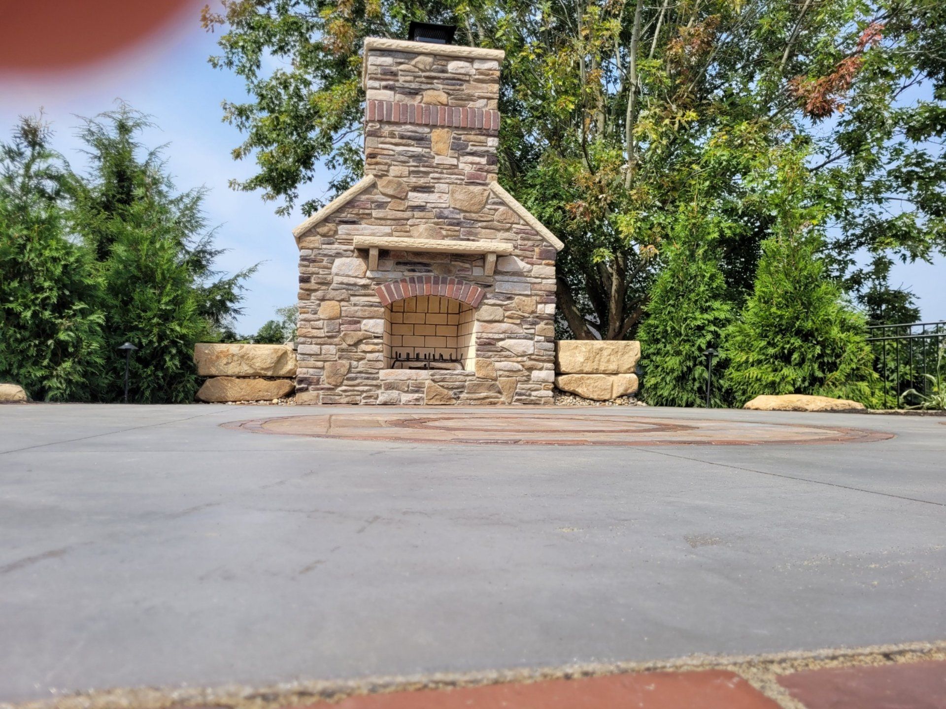 Stone outdoor fireplace with brick details and surrounding patio. Trees in background.