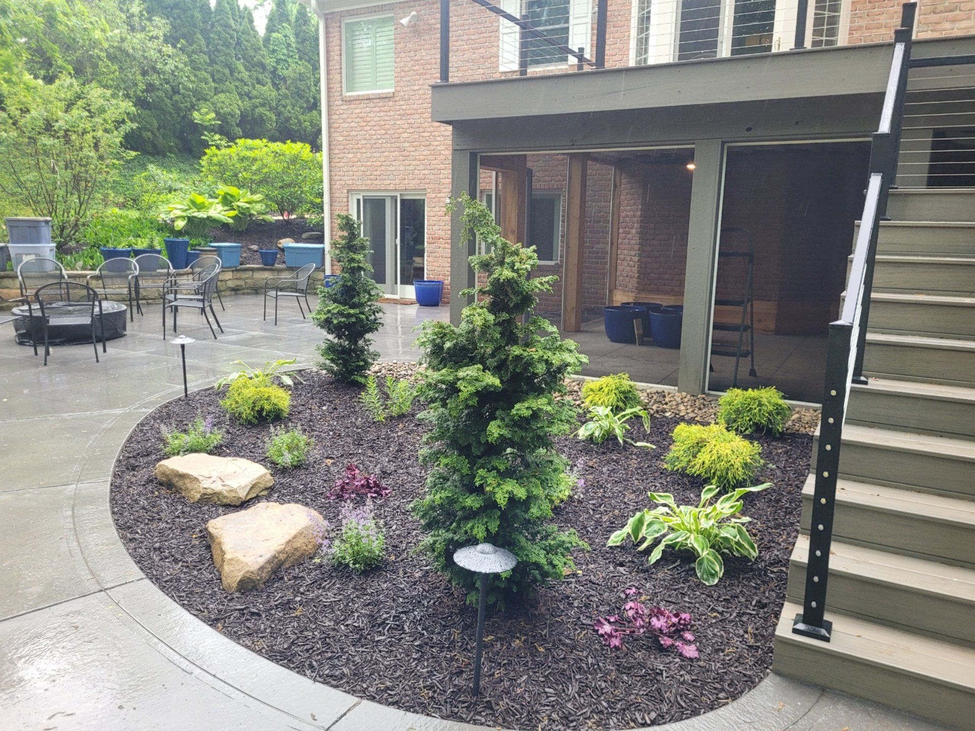 Landscaped backyard with trees, shrubs, and large rocks in a bed of dark mulch. Brick house and patio visible.