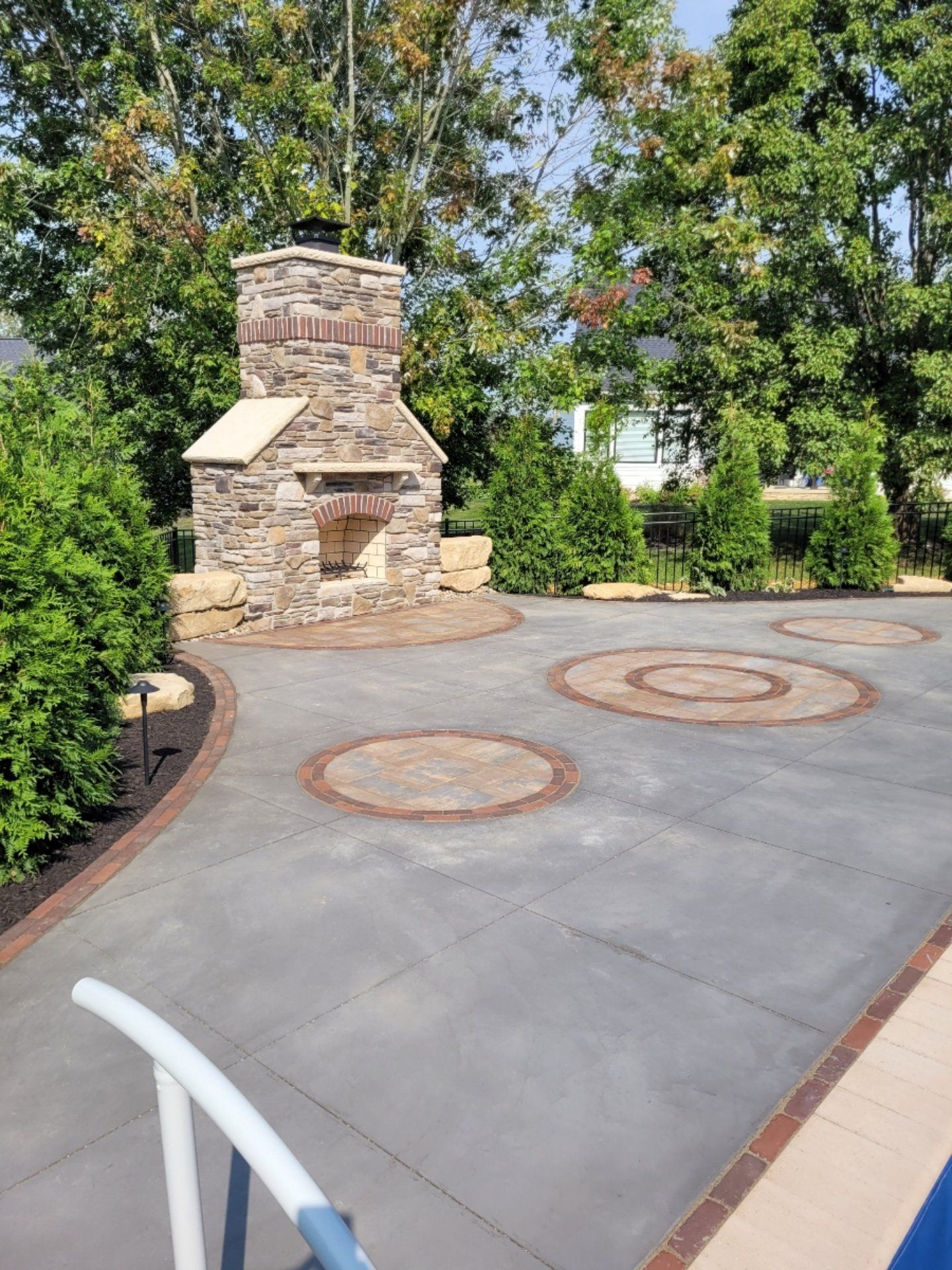 Outdoor stone fireplace and patio with circular brick and concrete patterns, surrounded by green trees and bushes.