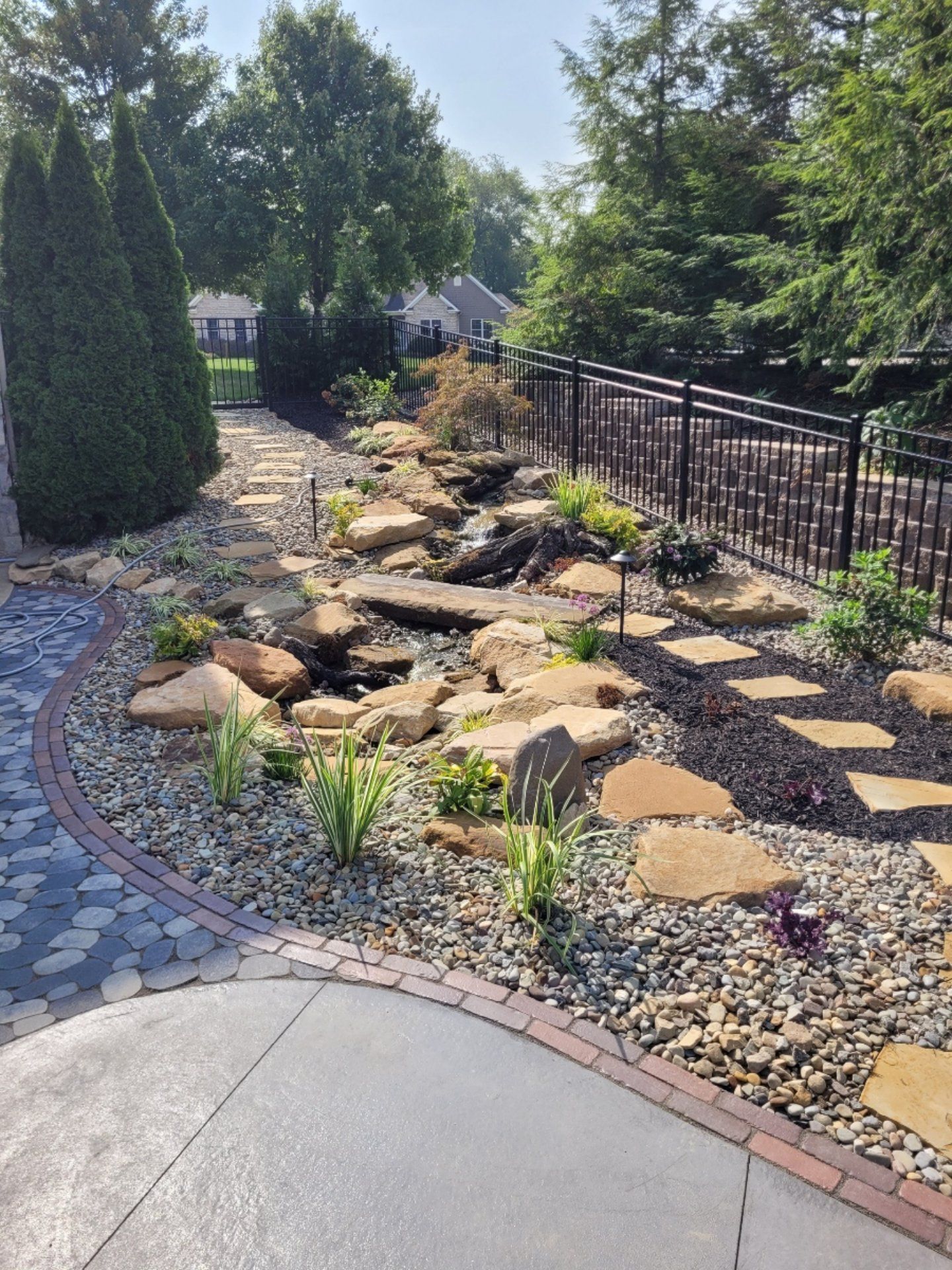 Stone pathway and water feature in a garden, with rocks, plants, and black fence.