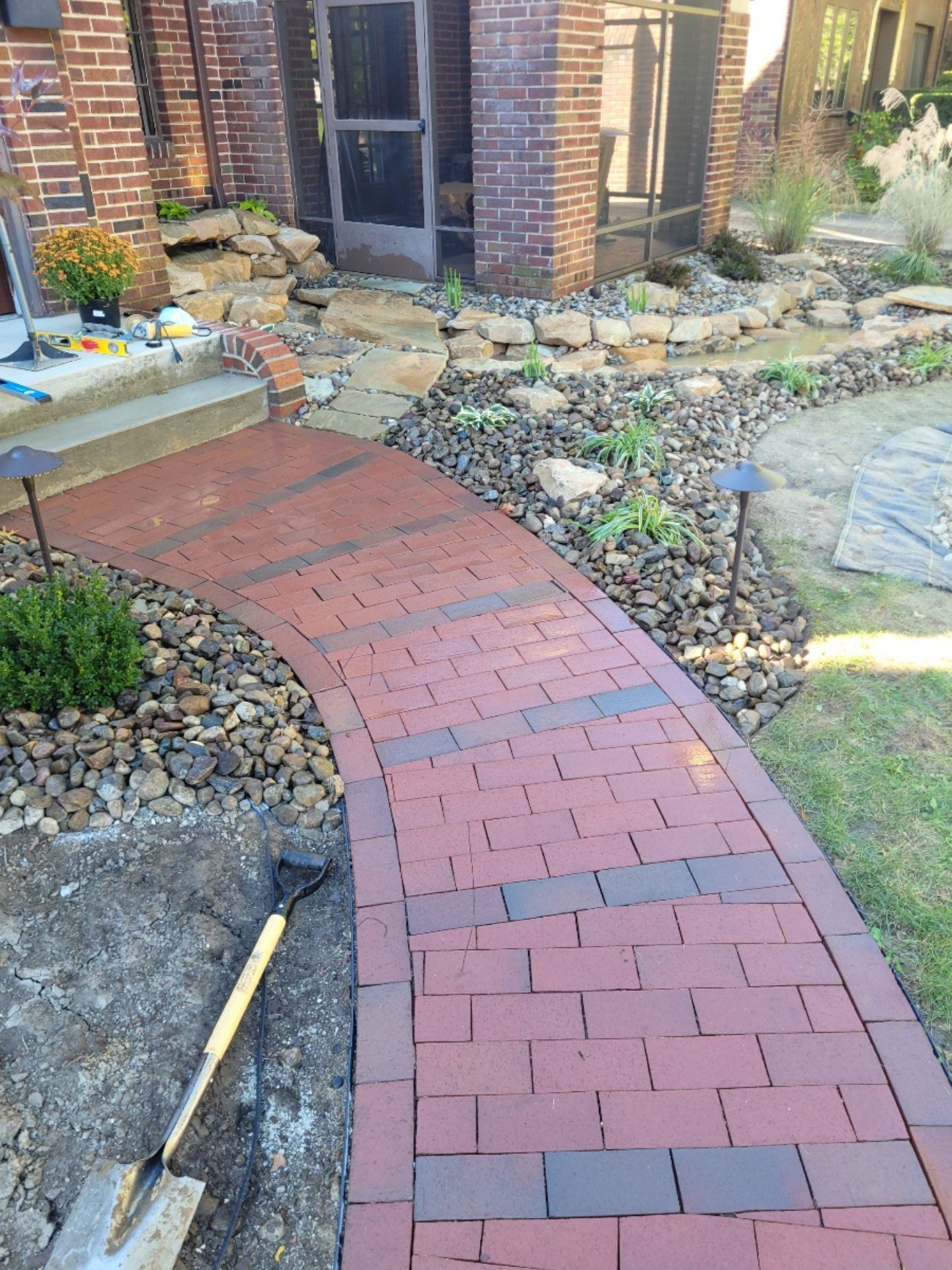 Brick pathway curves toward a doorway with landscaping and a shovel nearby.