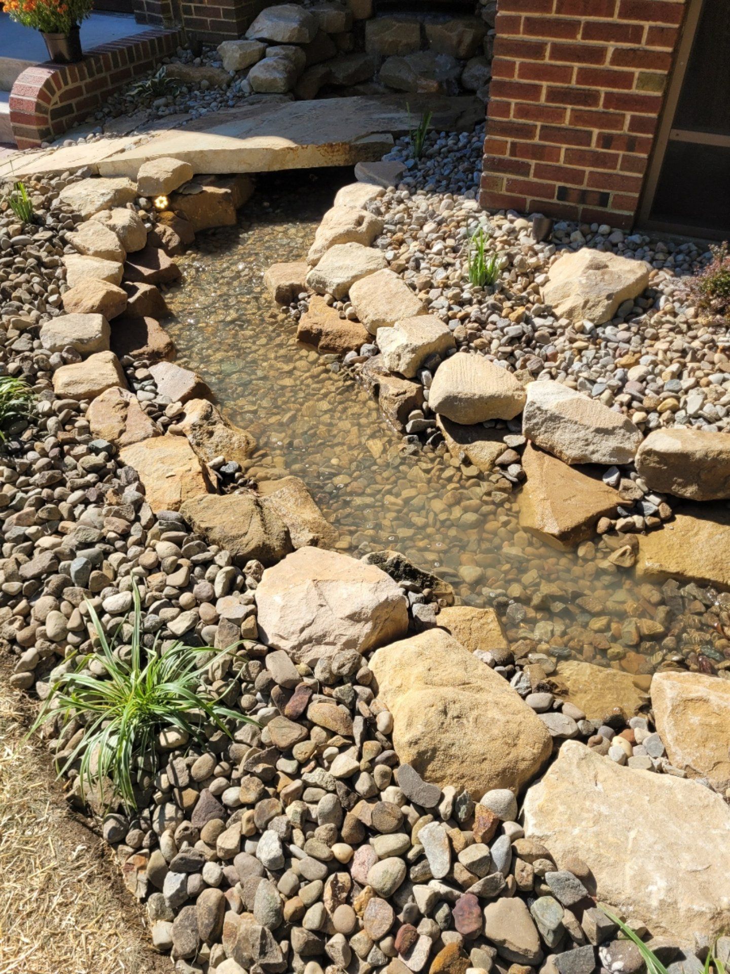A small water stream with rocks and stones runs alongside a brick building, with a stone bridge above.