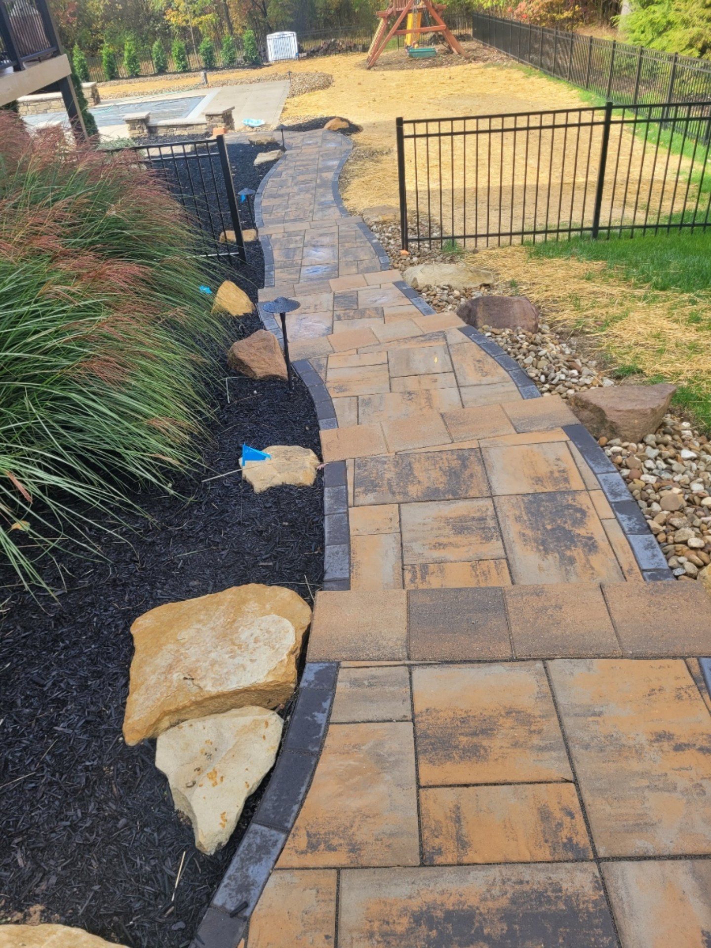 Stone pathway with steps in a backyard, bordered by black mulch, greenery, and large rocks.