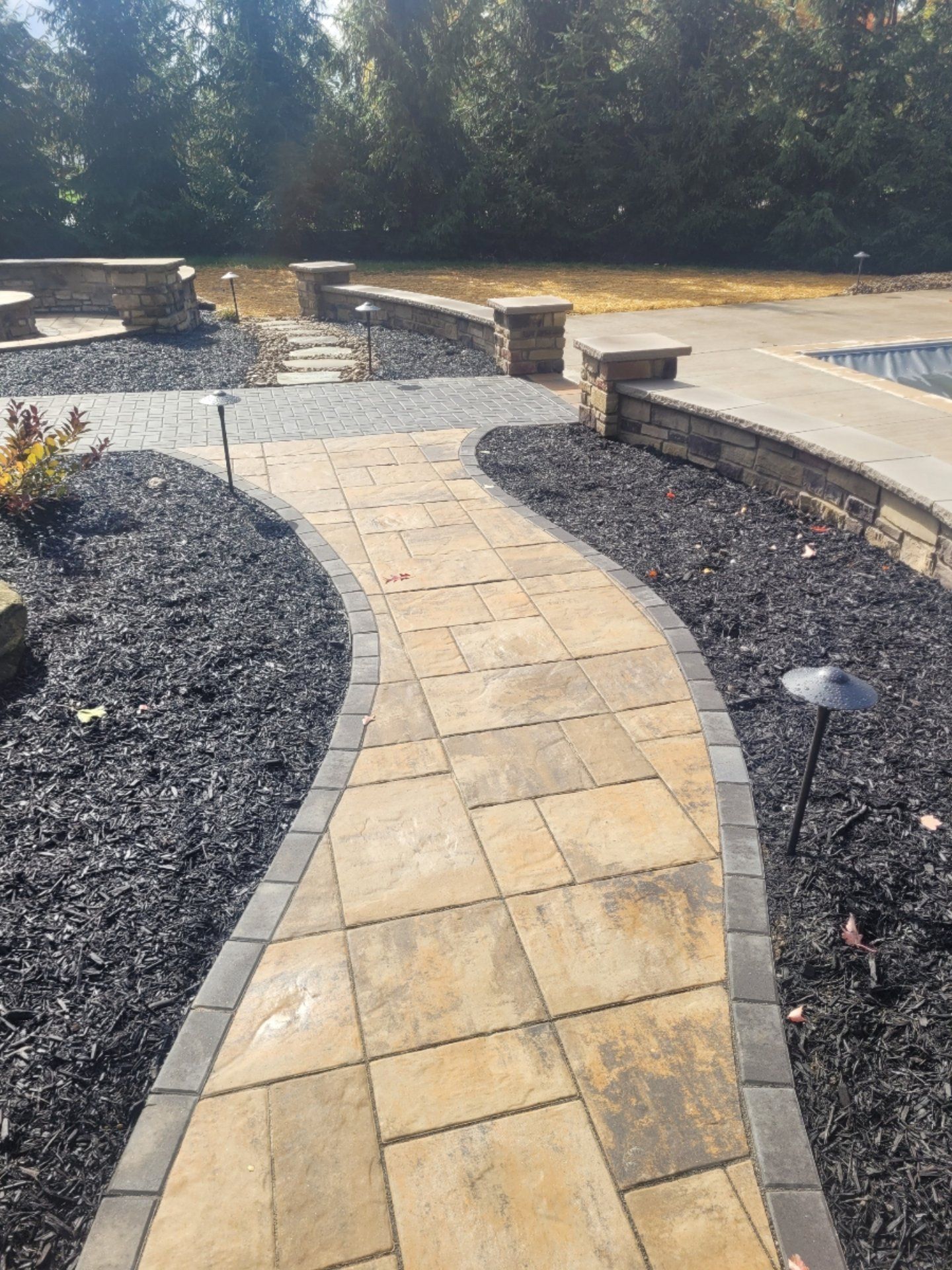 Stone pathway curves through black mulch, leading to a pool area with low walls.