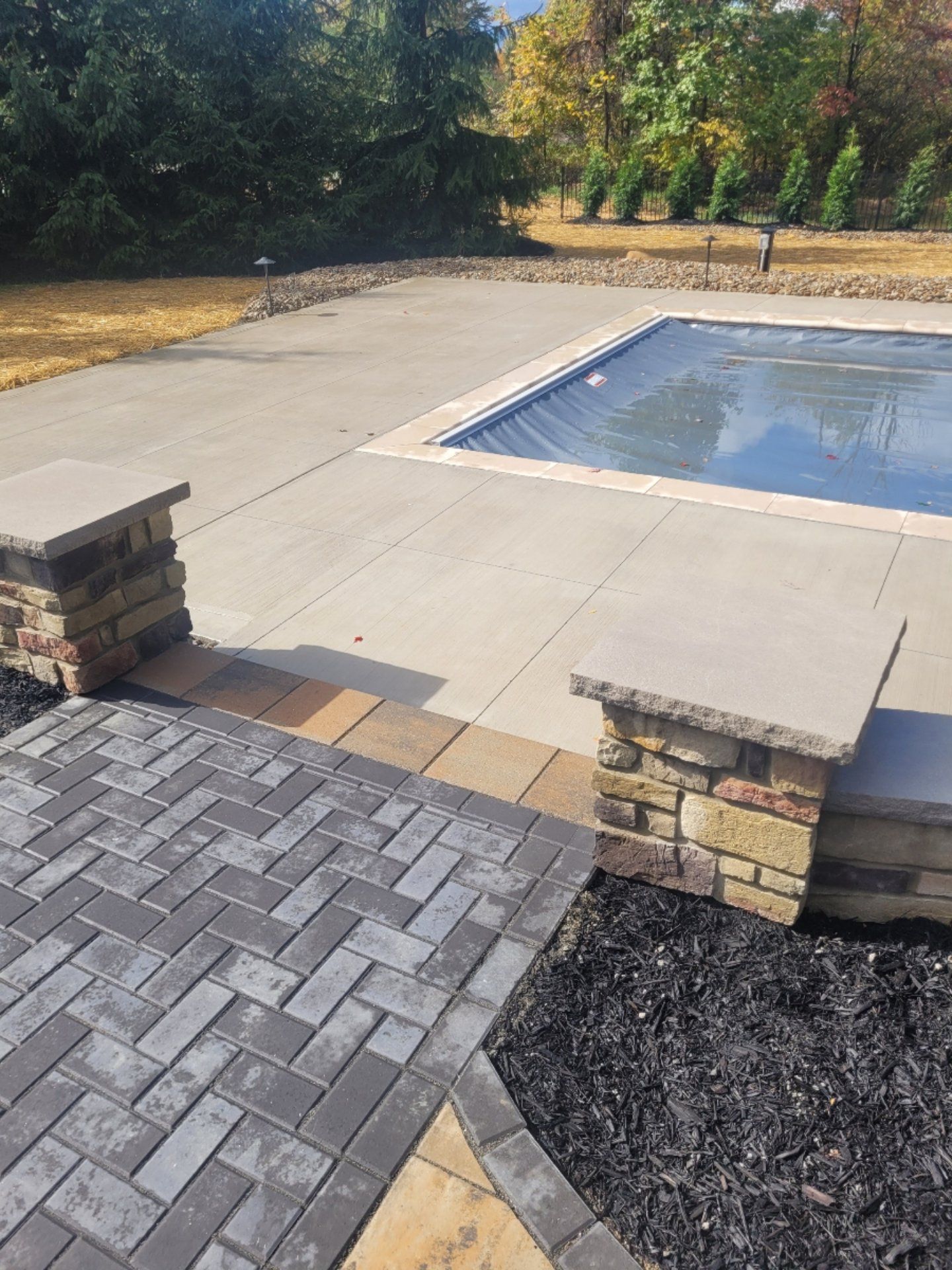 Concrete patio with a pool and brick pathway. Dark pool cover, brick columns, and a garden bed.