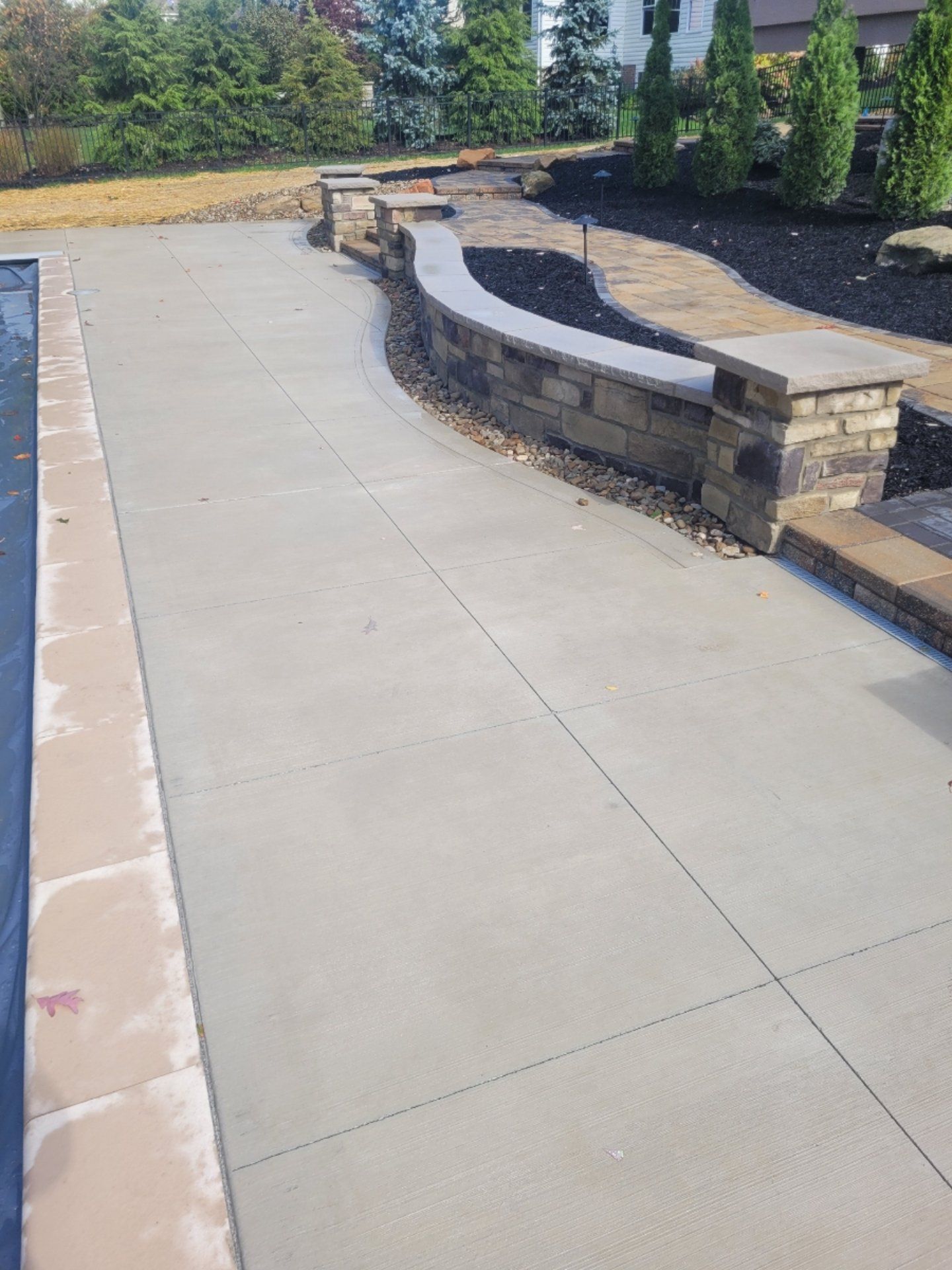 Concrete patio with a curved stone wall and pool border in a landscaped yard.