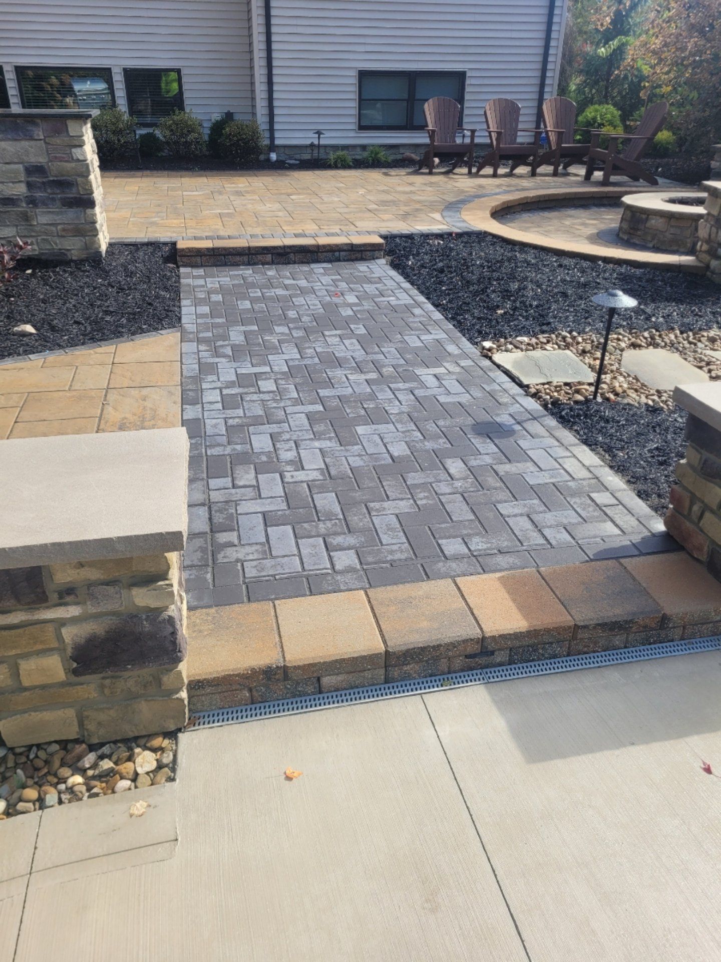 Brick pathway leading to a backyard patio with a fire pit, bordered by dark rocks and stone columns.