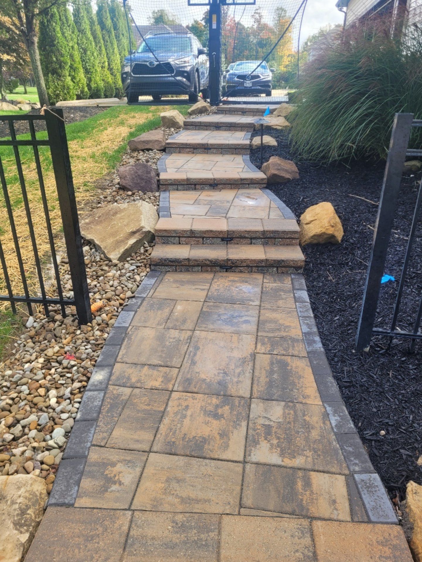 Paved walkway and steps leading to a driveway, bordered by rocks, black mulch, and a black fence.