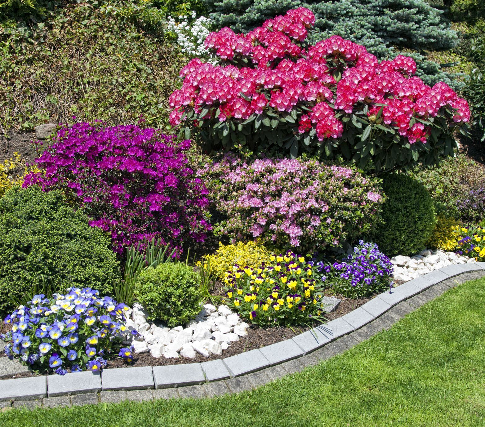 Colorful garden bed with blooming pink, purple, and yellow flowers, bordered by stone and white pebbles, next to green grass.