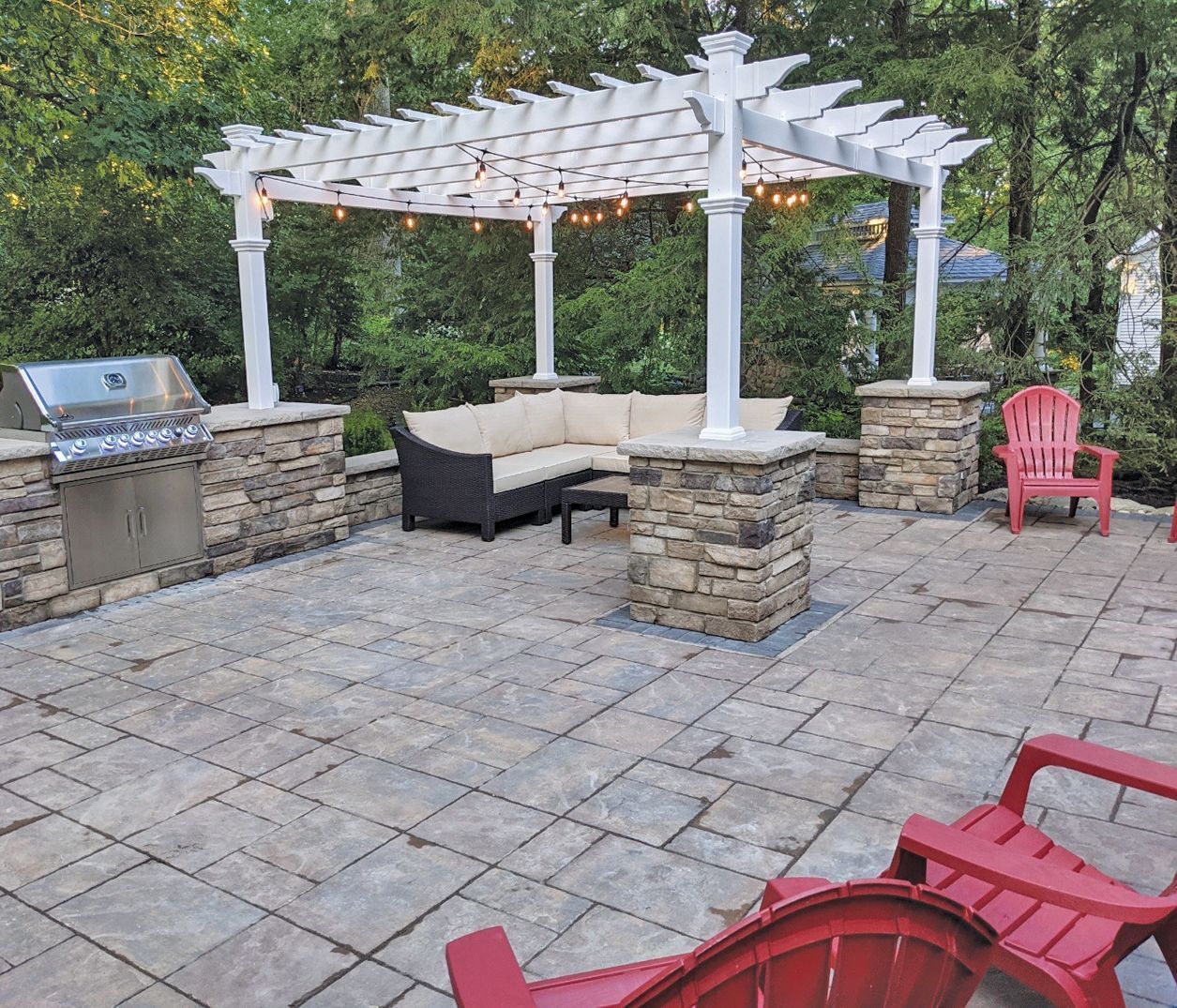 Outdoor patio with stone pavers, grill, seating, and a white pergola with string lights.  Red Adirondack chairs are present.