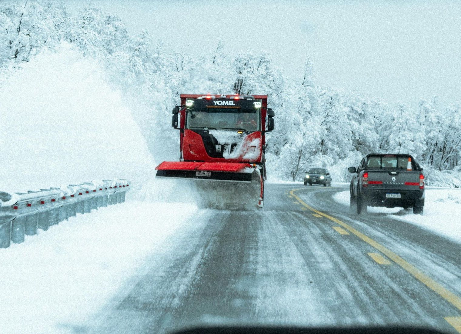 Snowplow clearing a snowy road; two cars follow.