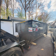 Black dump trailer parked on grass beside a brick house with a side staircase.