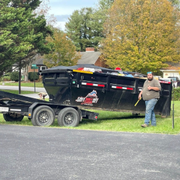 Man standing beside a black dump trailer on a grassy roadside in a suburban neighborhood