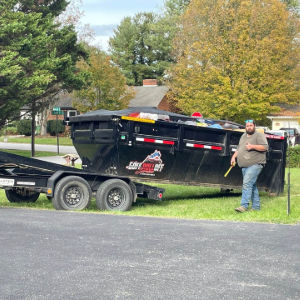 Man standing beside a black dump trailer on a grassy roadside in a suburban neighborhood