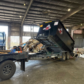 Dump trailer tipped up in an indoor warehouse, spilling debris onto the floor next to a flatbed truck.