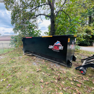 Black dumpster trailer on grass under trees, with a logo on the side and another trailer partly visible.
