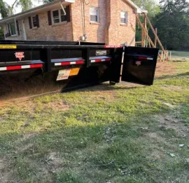 Black dump trailer in a narrow driveway beside parked vehicles and a white building under trees.