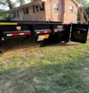 Black utility dumpster on a driveway between a white garage and a parked truck, with trees overhead.