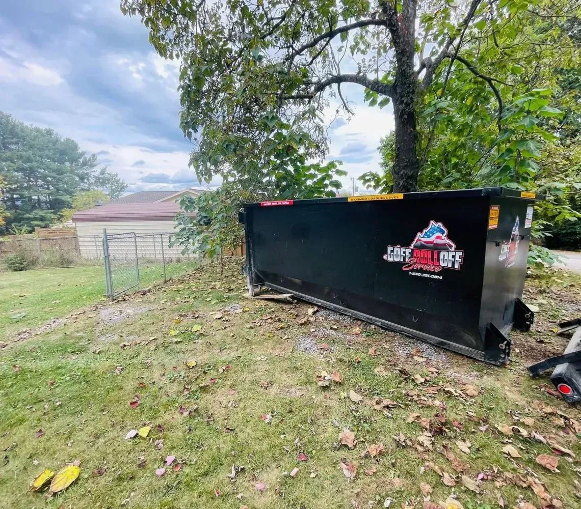 Black dumpster in a leafy yard with a logo on the side and a house in the background