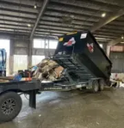 Dump trailer tipping over indoors with debris spilling onto the floor, beside a truck.