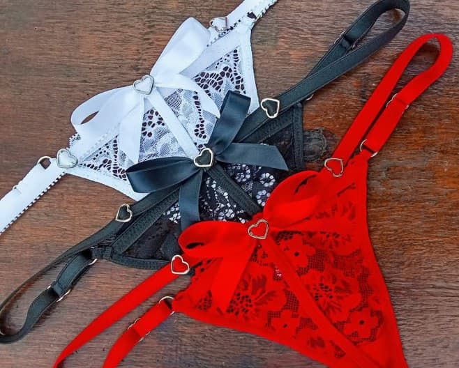 Ropa interior tipo tanga de encaje rojo y negro con lazos y detalles en forma de corazón, sobre fondo gris.