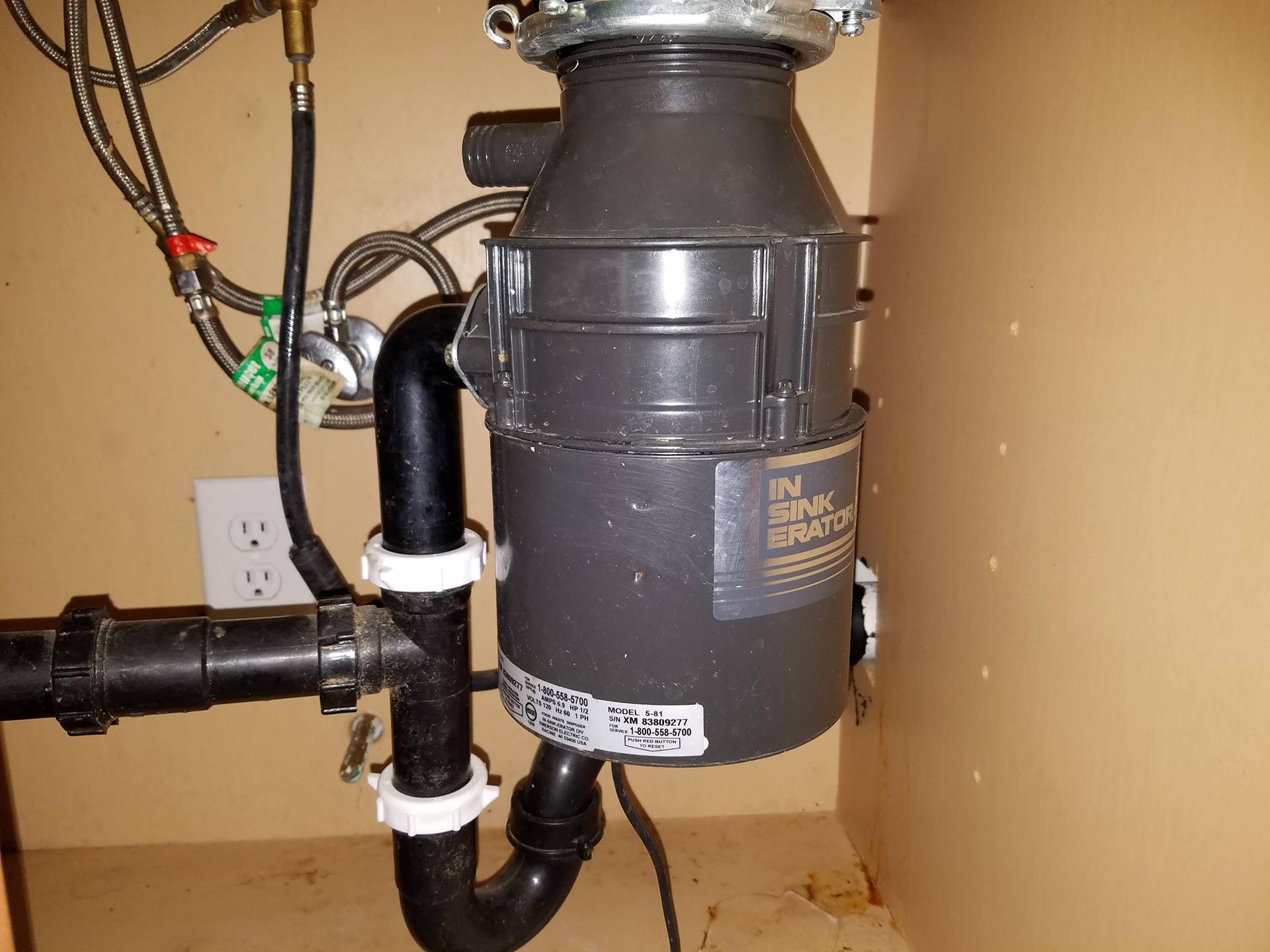 Under-sink garbage disposal and plumbing pipes against a beige wall.