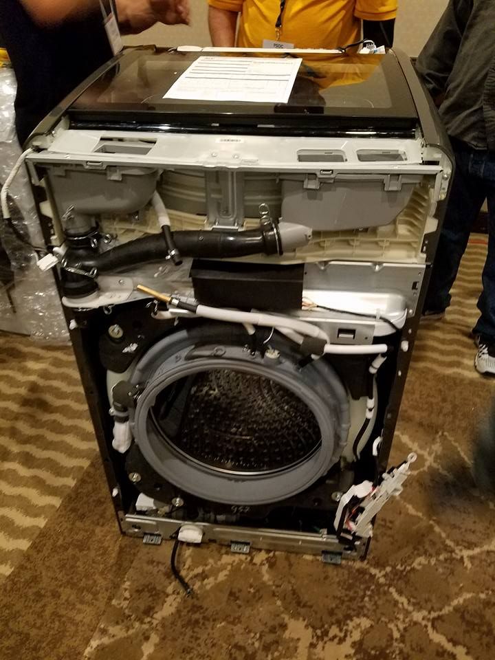 Broken front-loading washer with its front panel removed, exposing internal parts on a tiled floor.