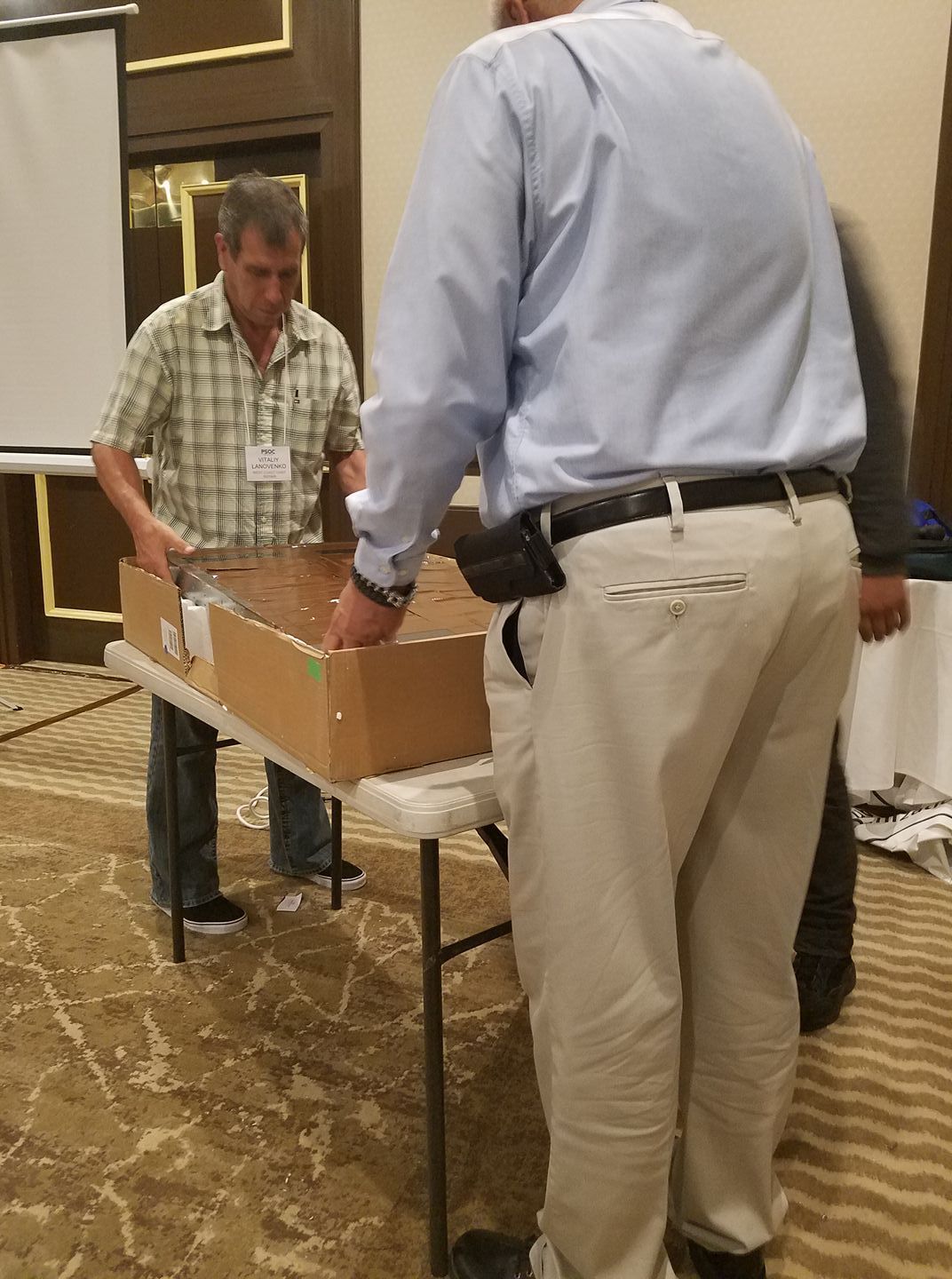 Two men unpacking a box at a conference table in a hotel hallway