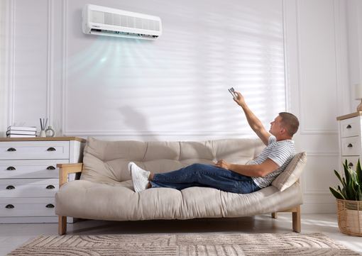 A person relaxing on a beige sofa in a bright room, using a remote control to adjust a wall-mounted air conditioner.
