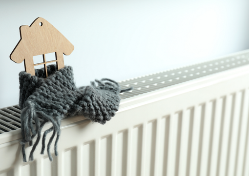 A small wooden house cutout wrapped in a thick, gray knitted scarf sits on top of a white radiator.