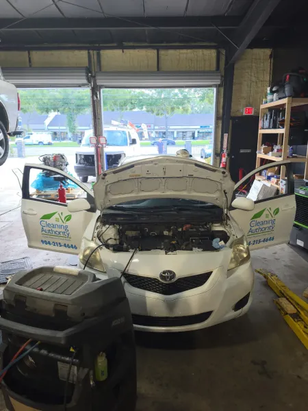 White Toyota sedan in a garage with hood open, surrounded by equipment and shelves during car repair
