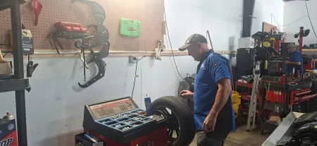 Man in blue shirt working in a cluttered auto repair garage beside a car wheel