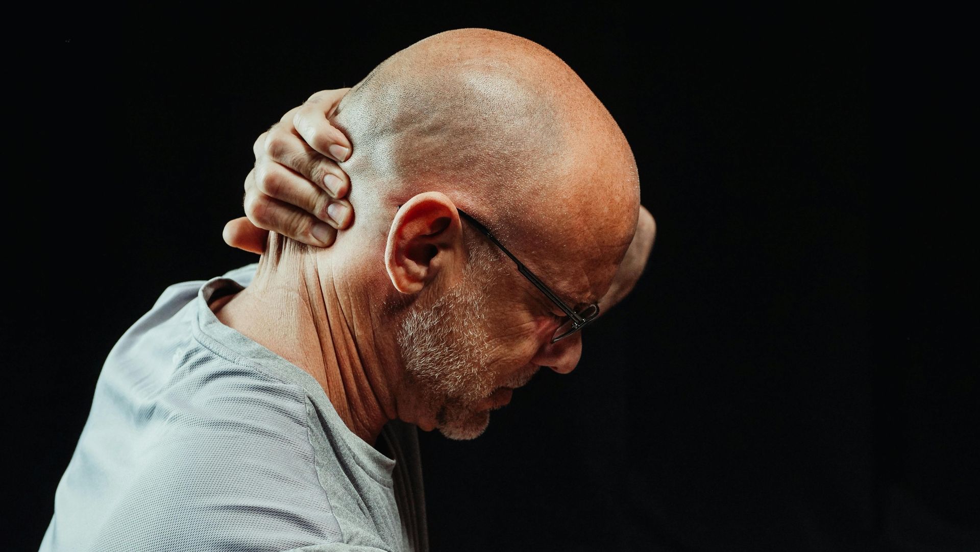 Man holding the back of his neck in pain, wearing glasses and a gray shirt against a black background