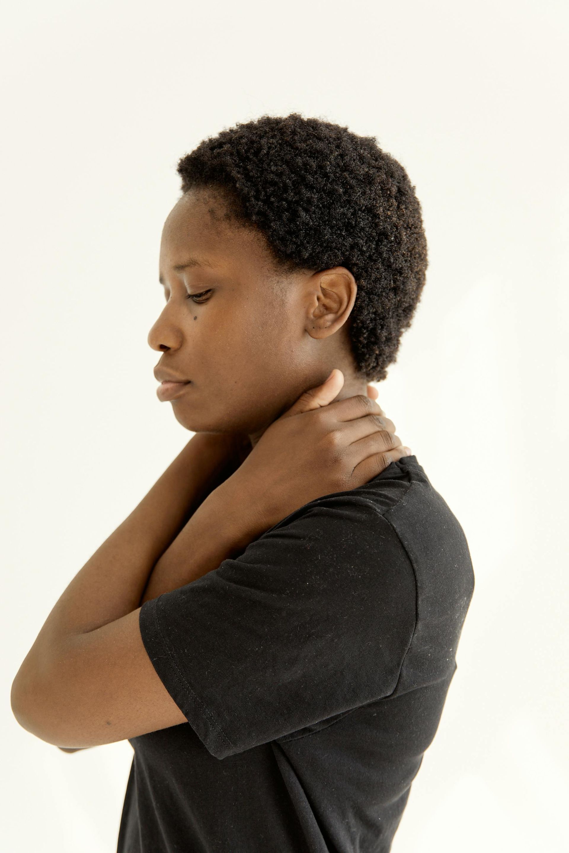 Person in a black shirt touching the back of their neck, looking down in a side profile against a plain background