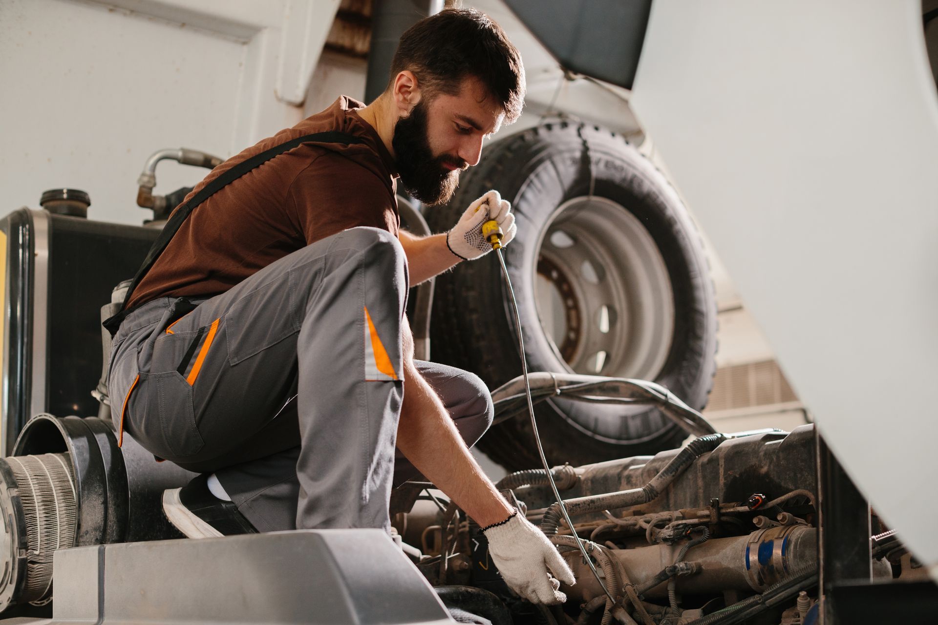Mechanic kneeling by a vehicle engine in a garage, holding a tool with tires in the background