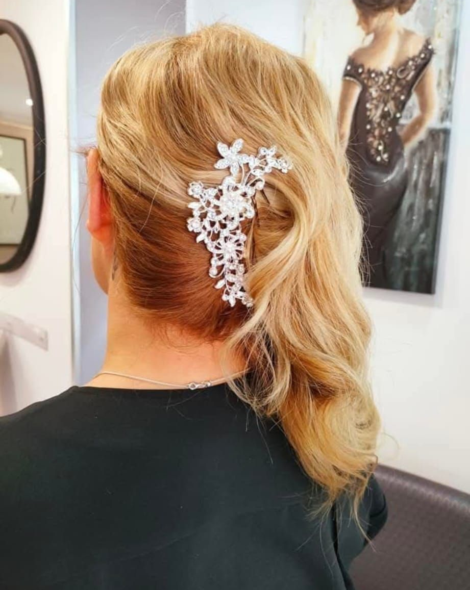 Una persona con i capelli biondi pettinati di lato, impreziositi da un fermaglio decorativo floreale bianco sul lato.