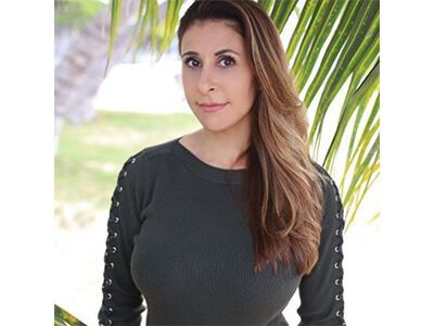 Woman in a dark top standing outdoors near palm leaves, looking at the camera.