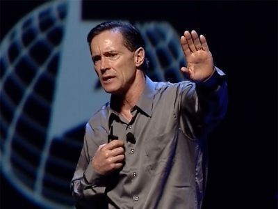 Man speaking on stage, holding a microphone and raising one hand, with a globe graphic behind him.