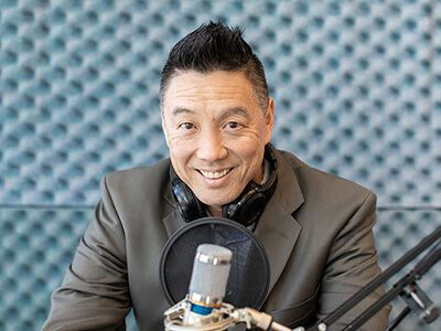Man smiling at a studio microphone, wearing a gray blazer against a blue acoustic wall.