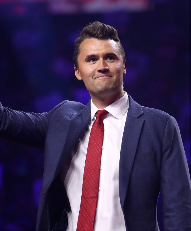 Man in a navy suit and red tie onstage, raising one arm against a purple-lit background